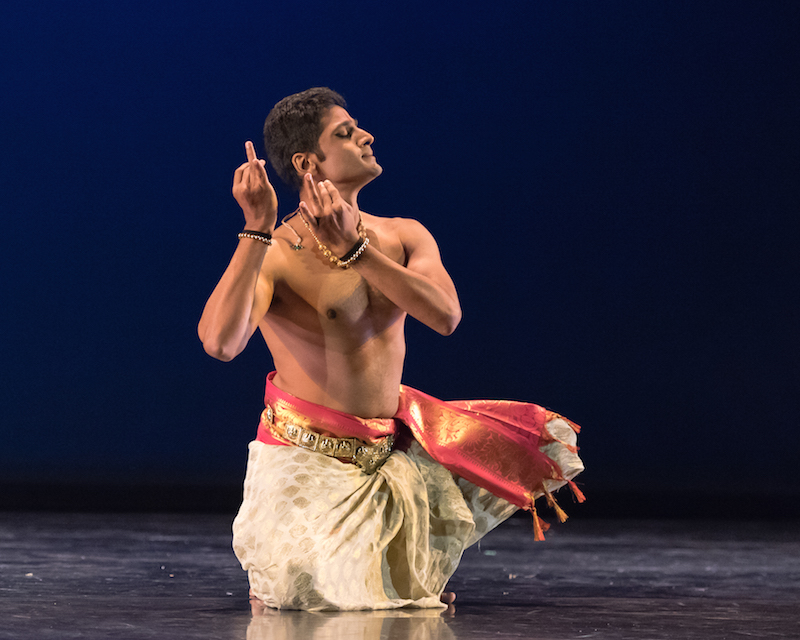 A bare-chested male Indian dancer assumes a kneeling position while his hands and fingers make a gesture near his face.